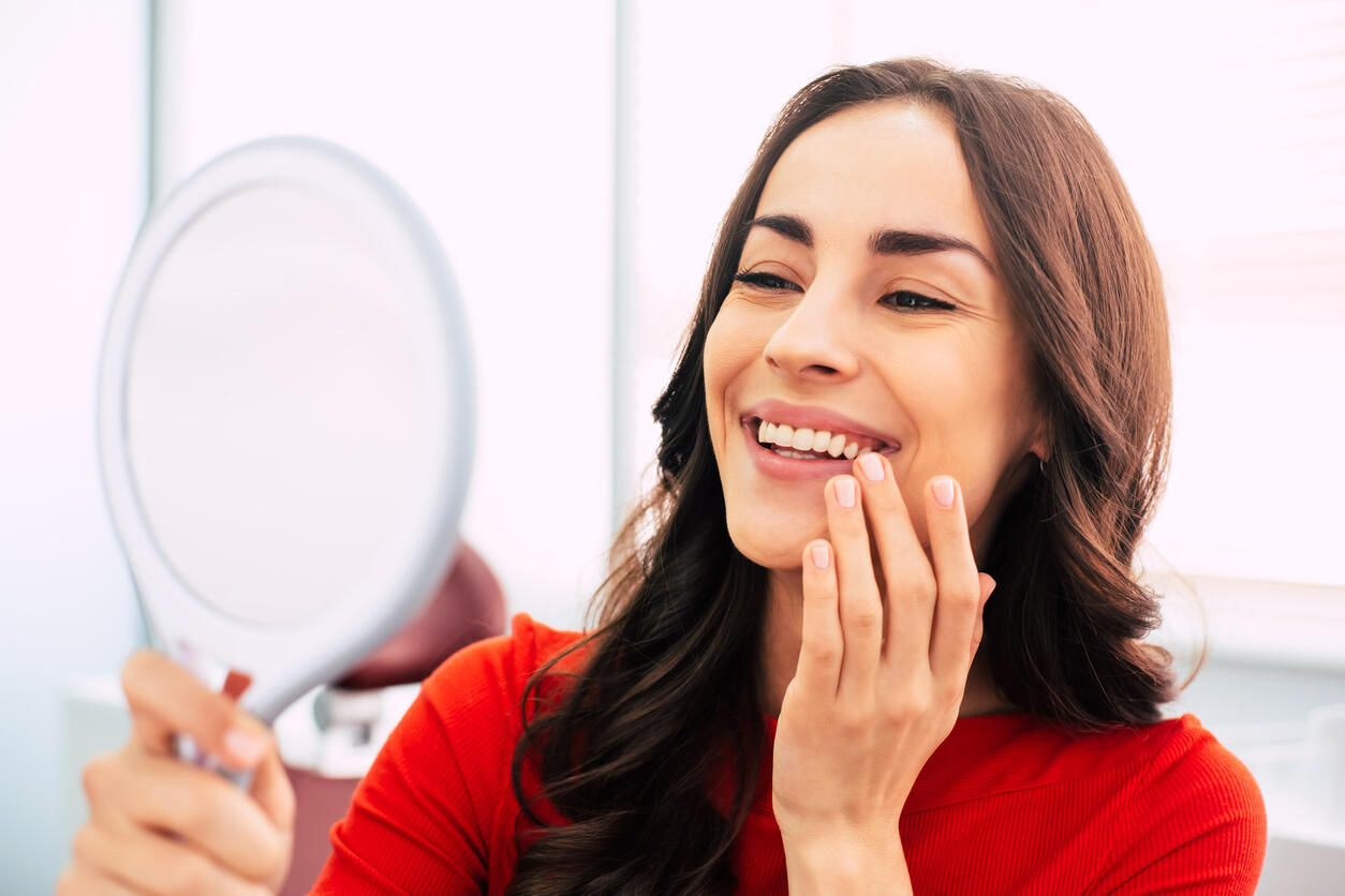 Honed to perfection. Fabulous woman clothed in red sweater, with curly long hair and elegant hands is holding a mirror for looking through the final work of dental doctor.